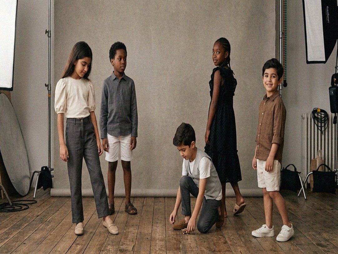 Children wearing relaxed, neutral-toned Ourson clothing in a daylit studio with a plaster backdrop and wooden floor.